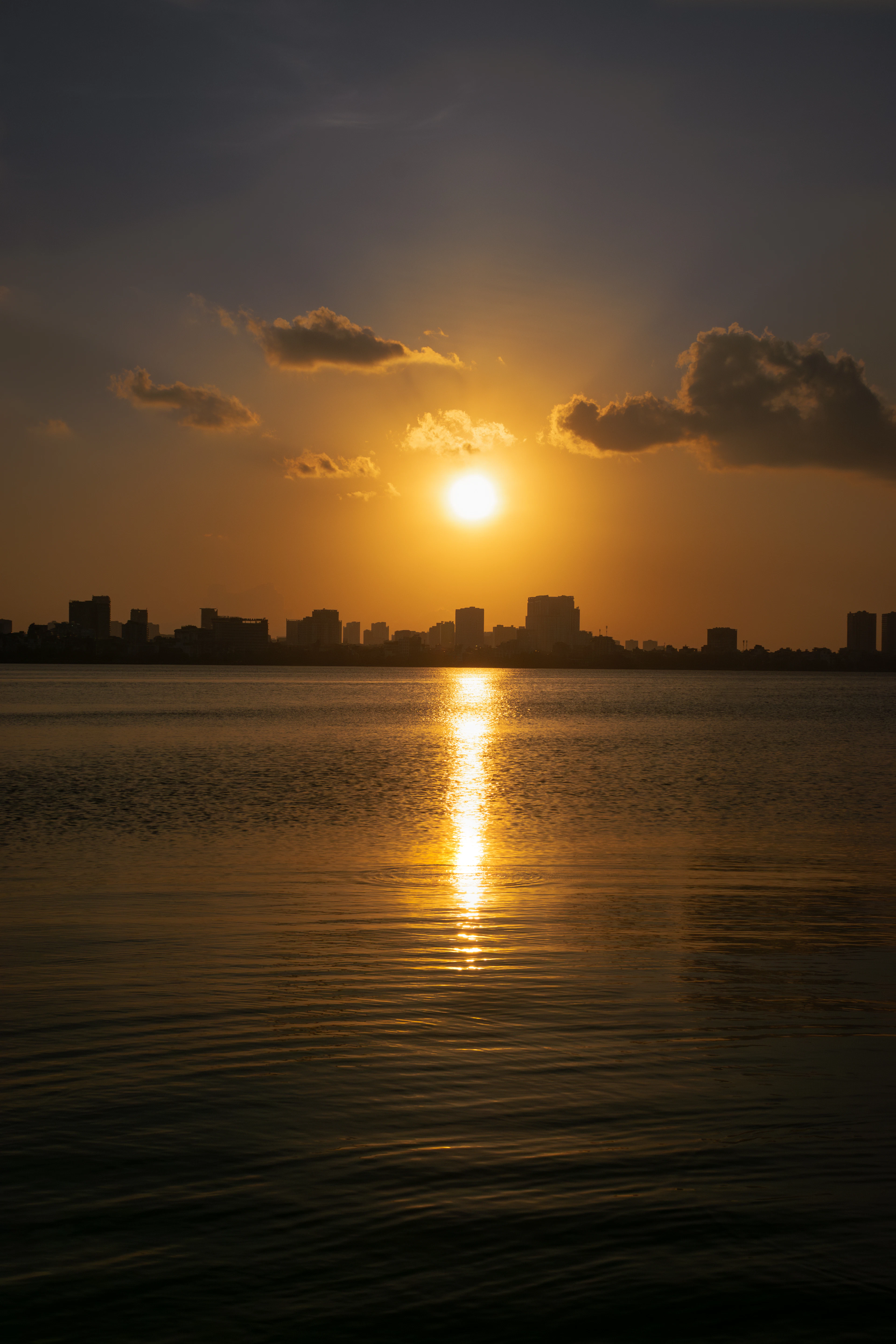 The sun setting over West Lake in Hanoi's Tay Ho district, a low orange disc between dark clouds above the silhouetted city skyline, laying a long column of gold light across the calm water.