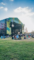 A large outdoor music stage set amidst a green grassy area with people sitting on the grass. The stage has vibrant lighting and colorful banners. The sky is clear with some clouds, creating a pleasant atmosphere.