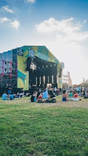 A large outdoor music stage set amidst a green grassy area with people sitting on the grass. The stage has vibrant lighting and colorful banners. The sky is clear with some clouds, creating a pleasant atmosphere.