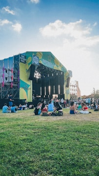 A large outdoor music stage set amidst a green grassy area with people sitting on the grass. The stage has vibrant lighting and colorful banners. The sky is clear with some clouds, creating a pleasant atmosphere.