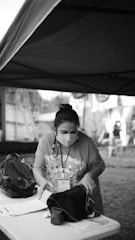 A person wearing a mask is organizing items on a table under a large canopy. The individual is focused on the contents of a bag placed on the table, which is also cluttered with papers. The setting seems to be outdoors, with other structures and people faintly visible in the background.
