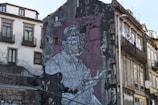 A large mural painted on the side of an urban building depicts an elderly person in a vest, wearing glasses, with an expressive gesture. The artwork is on a weathered wall with signs of decay, contrasting with adjacent buildings showcasing old European architecture with balconies.