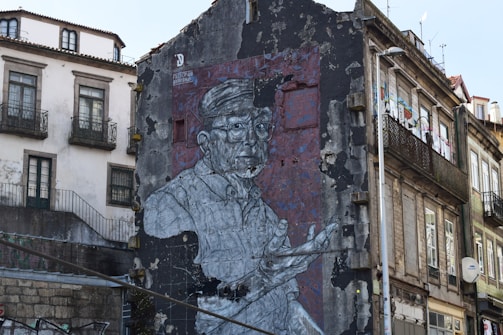A large mural painted on the side of an urban building depicts an elderly person in a vest, wearing glasses, with an expressive gesture. The artwork is on a weathered wall with signs of decay, contrasting with adjacent buildings showcasing old European architecture with balconies.
