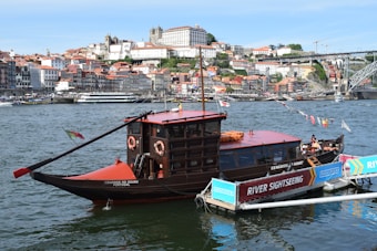 A scenic view featuring a traditional wooden boat used for river sightseeing, docked on a calm river. In the background, a historic city skyline with a mix of colorful buildings stretches across the hill, and a prominent bridge arches over the water. Flags decorate the side of the boat, adding vibrancy to the scene.
