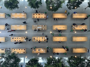 An overhead view of a large, modern store interior with several wooden tables arranged in rows. People are seen walking around or standing near the tables, some of which display electronics such as laptops and tablets. The store is spacious, with green trees interspersed among the tables, adding a touch of nature. The floor is light-colored, contributing to the clean and organized appearance of the space.