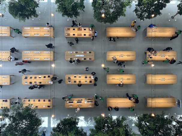 An overhead view of a large, modern store interior with several wooden tables arranged in rows. People are seen walking around or standing near the tables, some of which display electronics such as laptops and tablets. The store is spacious, with green trees interspersed among the tables, adding a touch of nature. The floor is light-colored, contributing to the clean and organized appearance of the space.