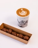 A cup of coffee with intricate latte art featuring a heart and leaf pattern on top, placed on a surface. Next to it, there is a cardboard box containing five round chocolates with a smooth exterior on display.