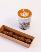 A cup of coffee with intricate latte art featuring a heart and leaf pattern on top, placed on a surface. Next to it, there is a cardboard box containing five round chocolates with a smooth exterior on display.