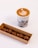 A cup of coffee with intricate latte art featuring a heart and leaf pattern on top, placed on a surface. Next to it, there is a cardboard box containing five round chocolates with a smooth exterior on display.