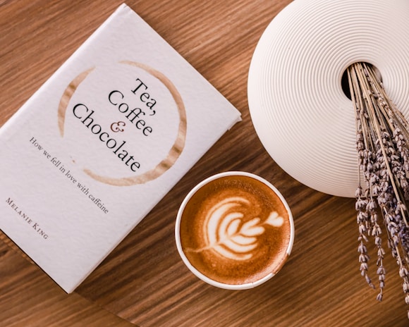 A book titled 'Tea, Coffee & Chocolate' is placed on a wooden table next to a cup of coffee with latte art on top. A decorative white vase filled with dried lavender stems is positioned on the side.