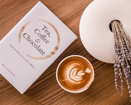 A book titled 'Tea, Coffee & Chocolate' is placed on a wooden table next to a cup of coffee with latte art on top. A decorative white vase filled with dried lavender stems is positioned on the side.