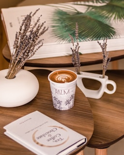 A cozy setup featuring a wooden table with a cup of coffee, beautifully topped with latte art. The cup is decorated with palm trees and the word 'PALM.' There is a white vase holding dried lavender, and a book titled 'Tea, Coffee & Chocolate.' In the background, there is a large, open book with an image of a green palm leaf.