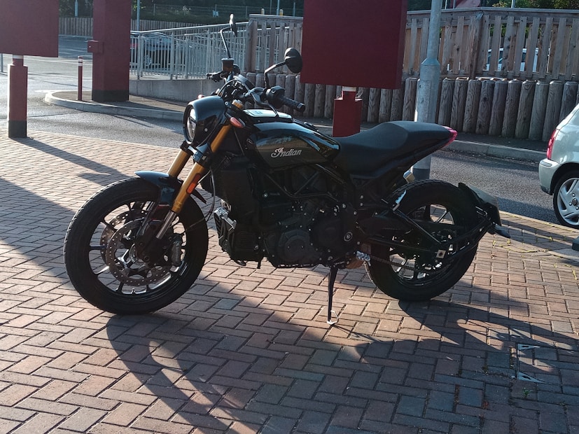 A sleek, black motorcycle parked on a sunlit brick pavement. The motorcycle features a sturdy frame, large wheels, and an overall modern design. It is situated near some fencing and an empty parking area, with visible shadows cast on the ground.