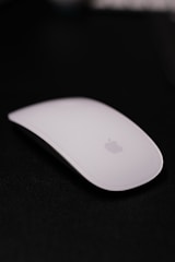 A sleek white computer mouse with a minimalist design placed against a dark background, featuring a subtle logo in the center.