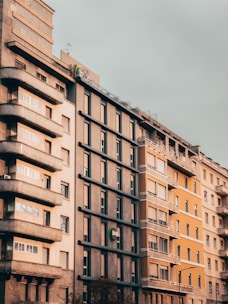 A row of multi-story urban buildings with a mix of architectural styles, featuring neutral and muted colors. The central building has a modern design with a logo on it, while the others have a more traditional appearance with balconies.