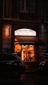 A warmly lit Chinese restaurant frontage at nighttime, displaying a sign that reads 'Rosticceria Cinese.' The interior showcases various decorations and wooden chairs, while a dark vehicle is parked in front.