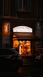 A warmly lit Chinese restaurant frontage at nighttime, displaying a sign that reads 'Rosticceria Cinese.' The interior showcases various decorations and wooden chairs, while a dark vehicle is parked in front.