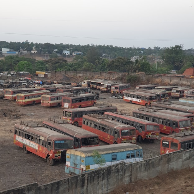 A large number of old, weathered buses are parked closely together in what appears to be a depot or scrapyard. The buses are predominantly red with some rust and signs of wear. The ground is a dusty, barren area, and a concrete wall surrounds the perimeter. In the background, there are trees and some distant buildings.