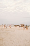 A group of people playing beach soccer on a sandy shoreline. In the background, there are beach huts with a clear sky overhead. The scene gives a casual and relaxed vibe as individuals engage in a leisurely outdoor activity.