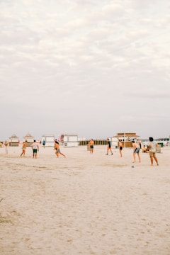 A group of people playing beach soccer on a sandy shoreline. In the background, there are beach huts with a clear sky overhead. The scene gives a casual and relaxed vibe as individuals engage in a leisurely outdoor activity.