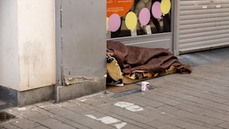 A person is lying on a mattress on the sidewalk next to a building. They are covered with a brown blanket. Nearby, there is an empty drink can placed on the ground. The area appears to be part of an urban street setting, with a closed shop featuring colorful circular signs in the window in the background.