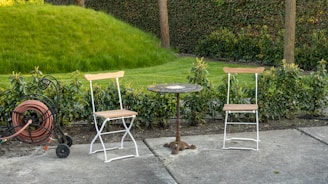 Two wooden chairs with metal frames are positioned on either side of a small, round table with a metal base. A garden hose is coiled on a reel nearby, and there is a lush green lawn with dense foliage in the background.