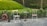 Two wooden chairs with metal frames are positioned on either side of a small, round table with a metal base. A garden hose is coiled on a reel nearby, and there is a lush green lawn with dense foliage in the background.