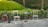 Two wooden chairs with metal frames are positioned on either side of a small, round table with a metal base. A garden hose is coiled on a reel nearby, and there is a lush green lawn with dense foliage in the background.