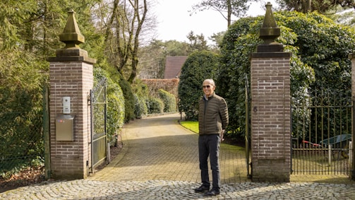 A man stands at the entrance of a driveway lined with tall hedges and trees. Two brick pillars with decorative tops frame the entrance, which is covered with a cobblestone path. A metal gate is attached to the right pillar. The background shows more greenery and a house with a pitched roof.