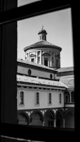 A black and white photograph showcasing a historical building with arched windows and a dome. The image is taken through a window, framing the architecture artfully.