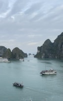 A scenic view of a serene bay featuring several boats on calm turquoise waters, surrounded by towering, lush green limestone islands under a cloudy sky.