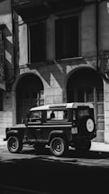 A vintage Land Rover is parked on a street in front of a building with arched doorways and large windows. The image is in black and white, creating dramatic shadows and highlights on the facade and vehicle.