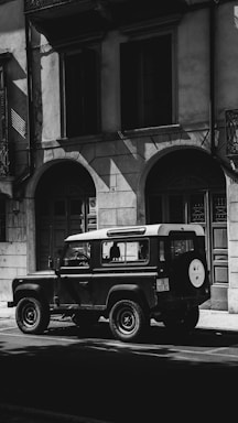 A vintage Land Rover is parked on a street in front of a building with arched doorways and large windows. The image is in black and white, creating dramatic shadows and highlights on the facade and vehicle.