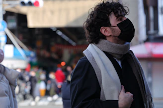 A person with curly hair wearing a black face mask and a gray scarf gazes upwards in an outdoor market setting. The background is bustling with blurred figures and market stalls, indicating a busy atmosphere.