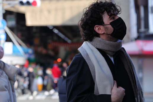 A person with curly hair wearing a black face mask and a gray scarf gazes upwards in an outdoor market setting. The background is bustling with blurred figures and market stalls, indicating a busy atmosphere.