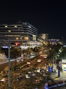 A night scene featuring a modern building with 'Conrad' signage on it, surrounded by streetlights and busy city traffic. The road is lined with trees and a parking area is visible on the right side. The atmosphere is lively with vehicles and people moving around.