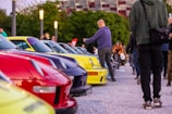 A vibrant outdoor automotive gathering with a row of colorful sports cars parked closely together. Several people are casually strolling and observing the vehicles while others are taking photos. The setting is in a public area with streetlights and a stadium visible in the background.