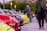 A vibrant outdoor automotive gathering with a row of colorful sports cars parked closely together. Several people are casually strolling and observing the vehicles while others are taking photos. The setting is in a public area with streetlights and a stadium visible in the background.