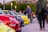 A vibrant outdoor automotive gathering with a row of colorful sports cars parked closely together. Several people are casually strolling and observing the vehicles while others are taking photos. The setting is in a public area with streetlights and a stadium visible in the background.
