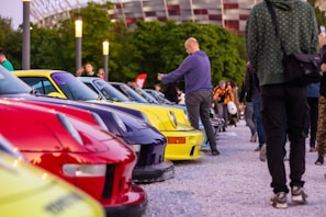 A vibrant outdoor automotive gathering with a row of colorful sports cars parked closely together. Several people are casually strolling and observing the vehicles while others are taking photos. The setting is in a public area with streetlights and a stadium visible in the background.