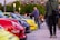 A vibrant outdoor automotive gathering with a row of colorful sports cars parked closely together. Several people are casually strolling and observing the vehicles while others are taking photos. The setting is in a public area with streetlights and a stadium visible in the background.