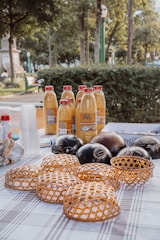 A table covered with a checkered cloth displays several bottles of orange juice with red caps, arranged neatly. In front of the bottles, there are decorative woven baskets with intricate patterns, along with black, round gourds featuring carved designs. In the background, a park setting is visible with green bushes, trees, and a monument.