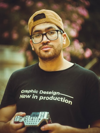 A man wearing a black t-shirt with the text 'Graphic Design — New in production' is holding a camera. He has a beige cap worn backwards and glasses. The background is blurred, with pink flowers and greenery.