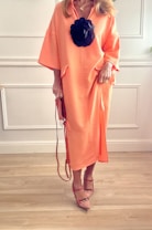 A person is wearing a long, flowing peach-colored dress with a large black floral accessory attached to the front. The outfit is complemented by strappy, pointed-toe shoes and a matching handbag, suggesting a fashion-forward and elegant style. The background features white paneling and wooden flooring, creating a clean and minimalist setting.