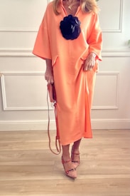 A person is wearing a long, flowing peach-colored dress with a large black floral accessory attached to the front. The outfit is complemented by strappy, pointed-toe shoes and a matching handbag, suggesting a fashion-forward and elegant style. The background features white paneling and wooden flooring, creating a clean and minimalist setting.