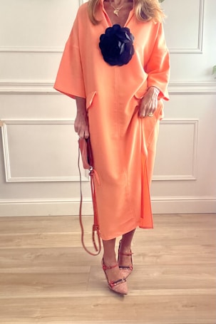 A person is wearing a long, flowing peach-colored dress with a large black floral accessory attached to the front. The outfit is complemented by strappy, pointed-toe shoes and a matching handbag, suggesting a fashion-forward and elegant style. The background features white paneling and wooden flooring, creating a clean and minimalist setting.