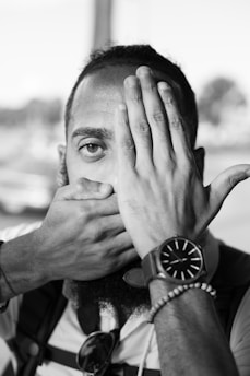 A person covers their mouth with one hand while the other hand covers half of their face. The individual is wearing a wristwatch and a bracelet on one wrist. The background is blurred, with indistinct shapes and light.