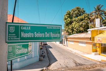 A street sign displaying 'Calle Nuestra Señora Del Rosario' is prominently positioned in an urban setting. The background consists of a sunny street lined with colorful buildings, including a yellow structure on the right. A tree provides some greenery amidst the urban landscape. Telephone lines are visible against a clear blue sky.