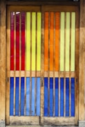 A set of wooden doors featuring vertical slats of colorful stained glass panels. The glass sections include rich hues of red, yellow, orange, and blue, creating a vibrant and eye-catching effect. The wooden frame has a natural finish, complementing the bright colors and adding warmth to the visual composition.