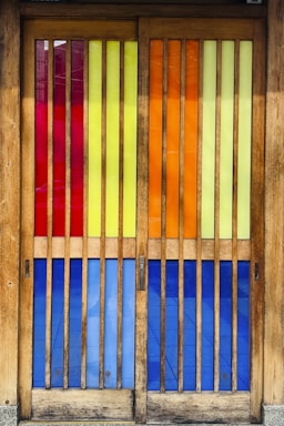 A set of wooden doors featuring vertical slats of colorful stained glass panels. The glass sections include rich hues of red, yellow, orange, and blue, creating a vibrant and eye-catching effect. The wooden frame has a natural finish, complementing the bright colors and adding warmth to the visual composition.